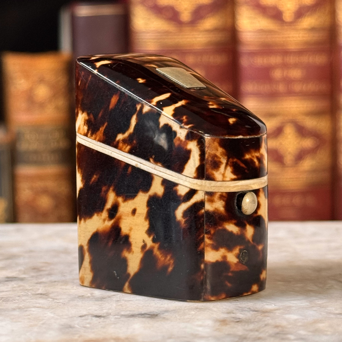 An antique tortoiseshell box with a glossy dark brown and amber pattern, featuring a gold-toned metal piece on top and a small round button, shown from side angle
