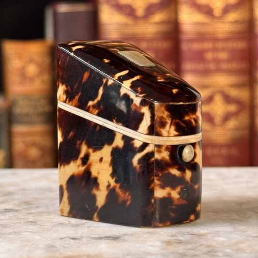 An antique tortoiseshell box with a glossy dark brown and amber pattern, featuring a gold-toned metal piece on top and a small round button, shown from side angle