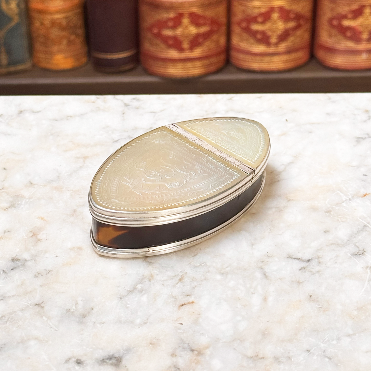 An antique oval tortoiseshell, mother-of-pearl, and silver snuff box, placed on a marble surface, angled overhead view