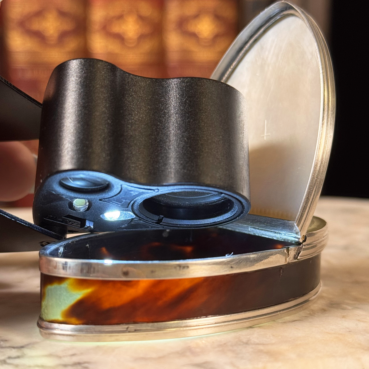 An antique oval tortoiseshell, mother-of-pearl, and silver snuff box, with a lighted loupe shining inside to highlight the tortoiseshell