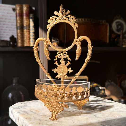 Decorative gold stand with a round center and glass dish on a marble surface. Features two swans, bows, and a cherub.