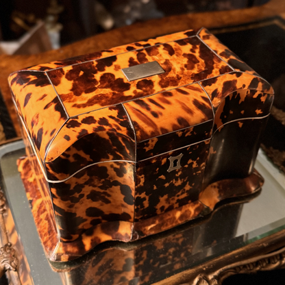 Antique Tortoiseshell Tea Caddy with curvature sitting on top of a gold mirror pedestal. The tea caddy has a silver escutcheon, and silver inlay. There is a small rectangle silver inlay on top of the hinged lid that can be engraved. Some professional repairs made to base edges. This is atop angled view.