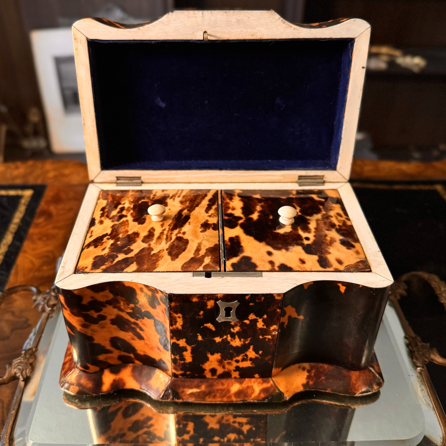 Antique Tortoiseshell Tea Caddy with curvature sitting on top of a gold mirror pedestal. The tea caddy is open and shows the two lids with small knobs. It also shows the blue velvet inside the upper lid. It has a silver escutcheon, and silver inlay. There is a small rectangle silver inlay on top of the hinged lid that can be engraved. Some professional repairs made to base edges. This is a front/top straight on view.