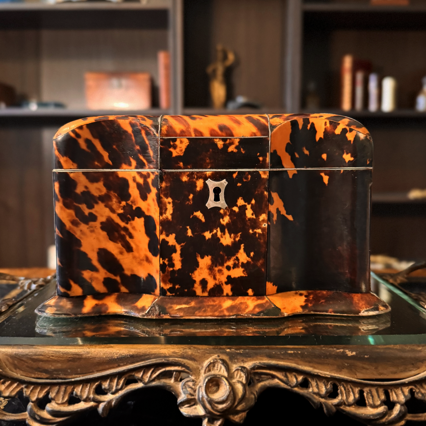 Antique Tortoiseshell Tea Caddy with curvature sitting on top of a gold mirror pedestal. The tea caddy has a silver escutcheon, and silver in lay. Some professional repairs made to base edges. This is a front view.